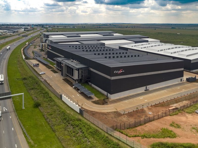 Aerial view of a large-scale industrial warehouse facility featuring extensive roofing, paved parking areas, and secure perimeter fencing adjacent to a major highway.