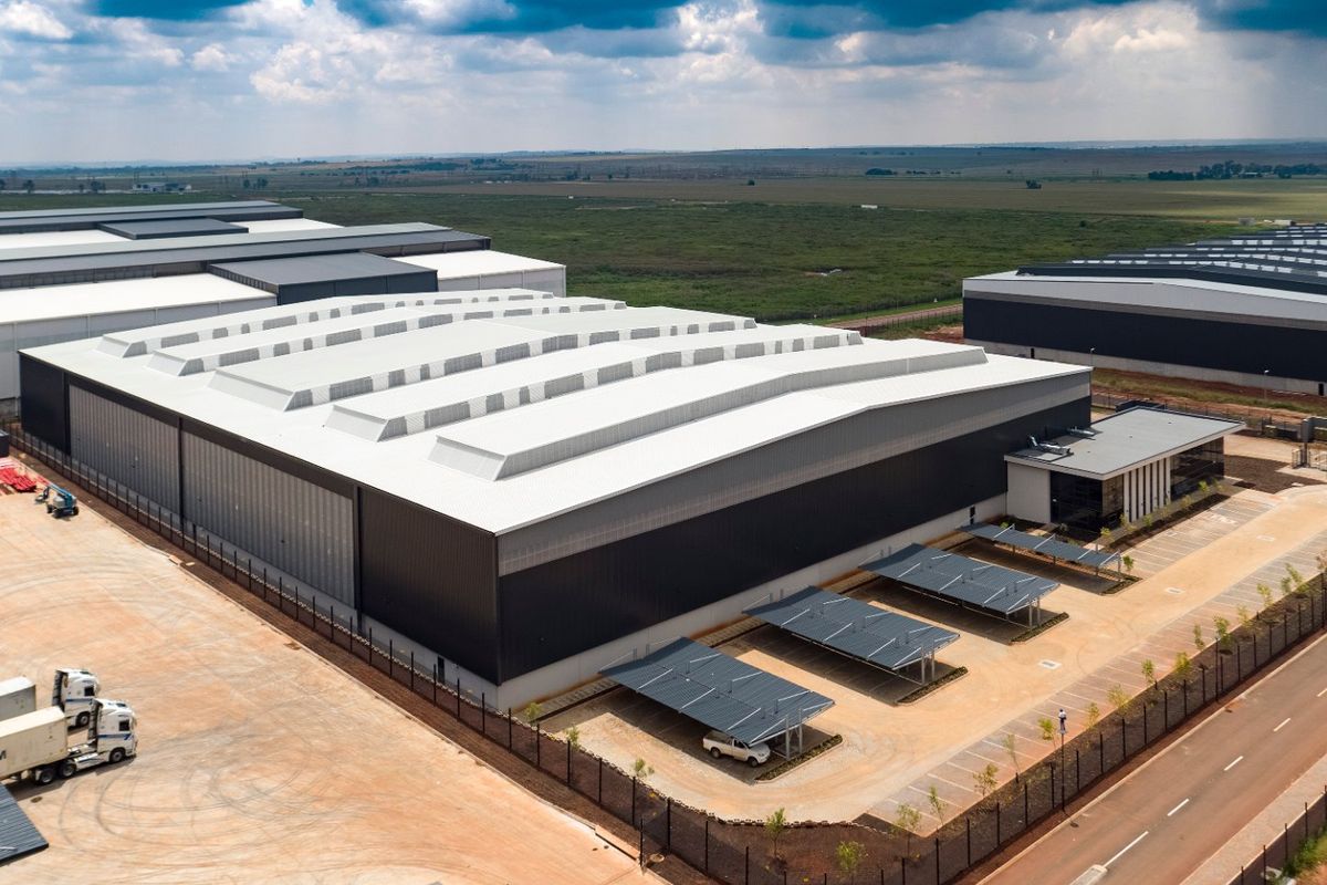 Aerial view of a modern industrial warehouse facility featuring a large metal roof, integrated skylights, paved parking area with carports, and secure perimeter fencing.
