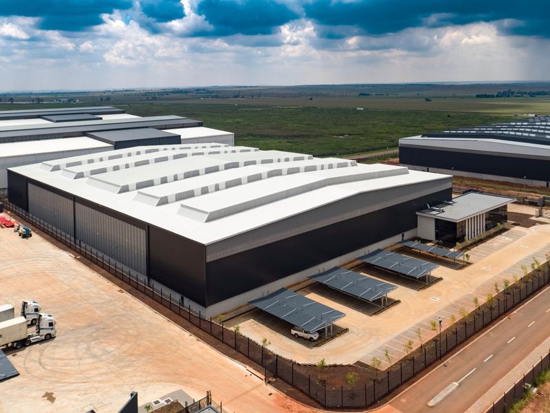 Aerial view of a modern industrial warehouse facility featuring a large metal roof, integrated skylights, paved parking area with carports, and secure perimeter fencing.