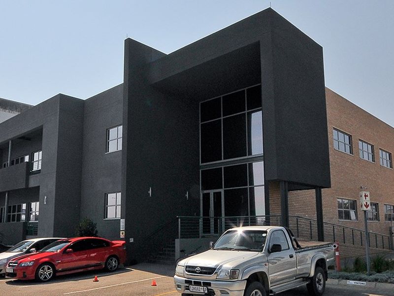 Modern industrial office and warehouse facility featuring a prominent glass entrance, dark textured facade, and dedicated paved parking area.