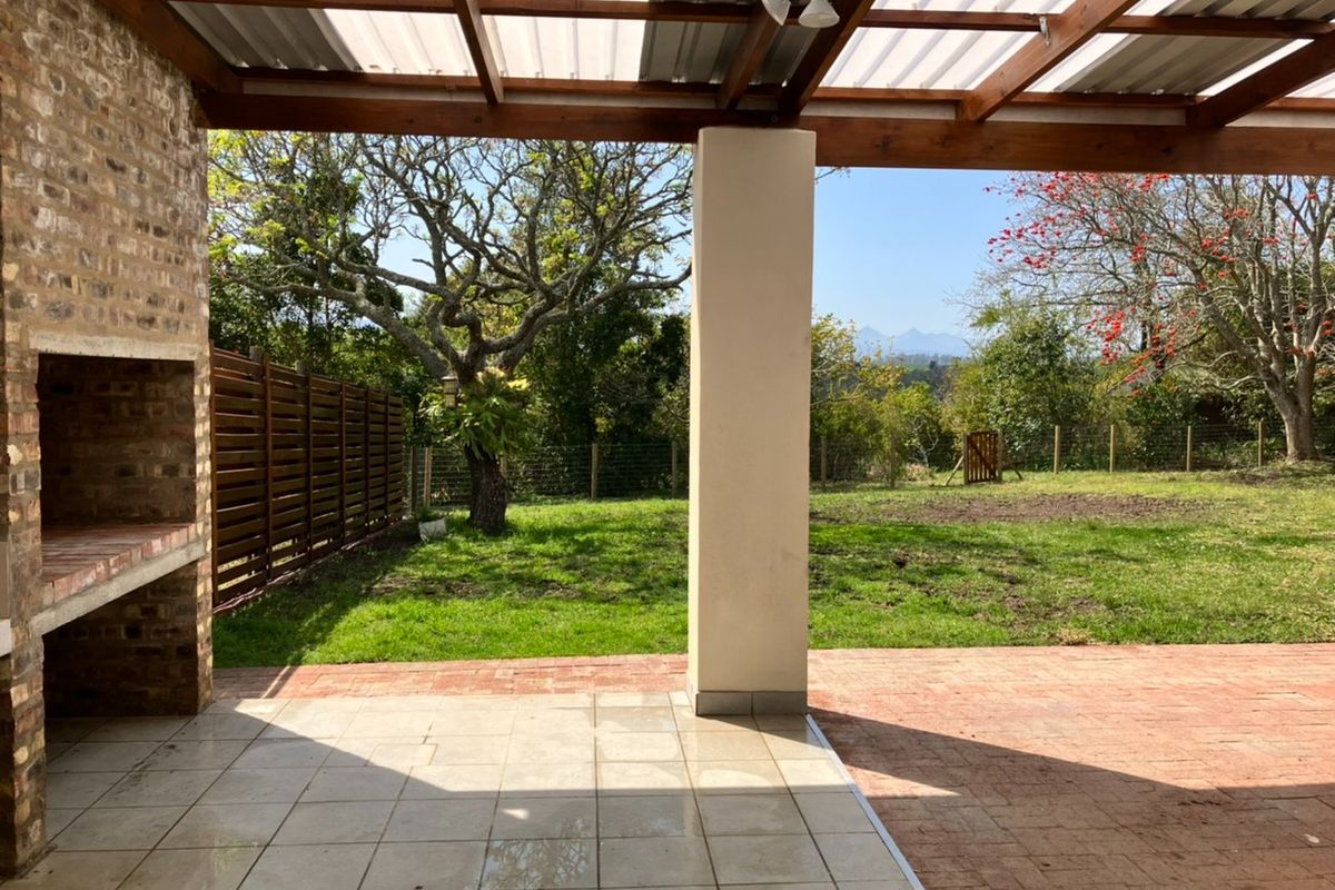 Covered outdoor patio featuring a built-in brick braai, timber pergola structure, and tiled flooring, overlooking a spacious garden with mountain views.