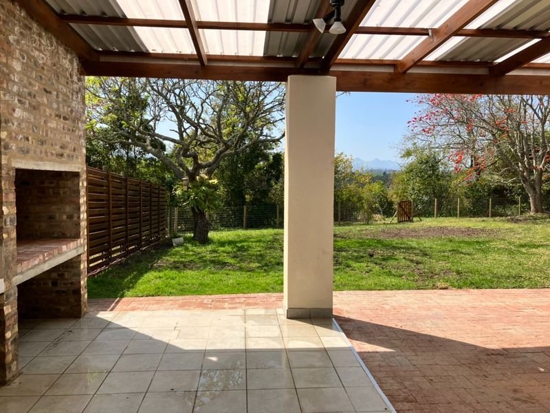 Covered outdoor patio featuring a built-in brick braai, timber pergola structure, and tiled flooring, overlooking a spacious garden with mountain views.