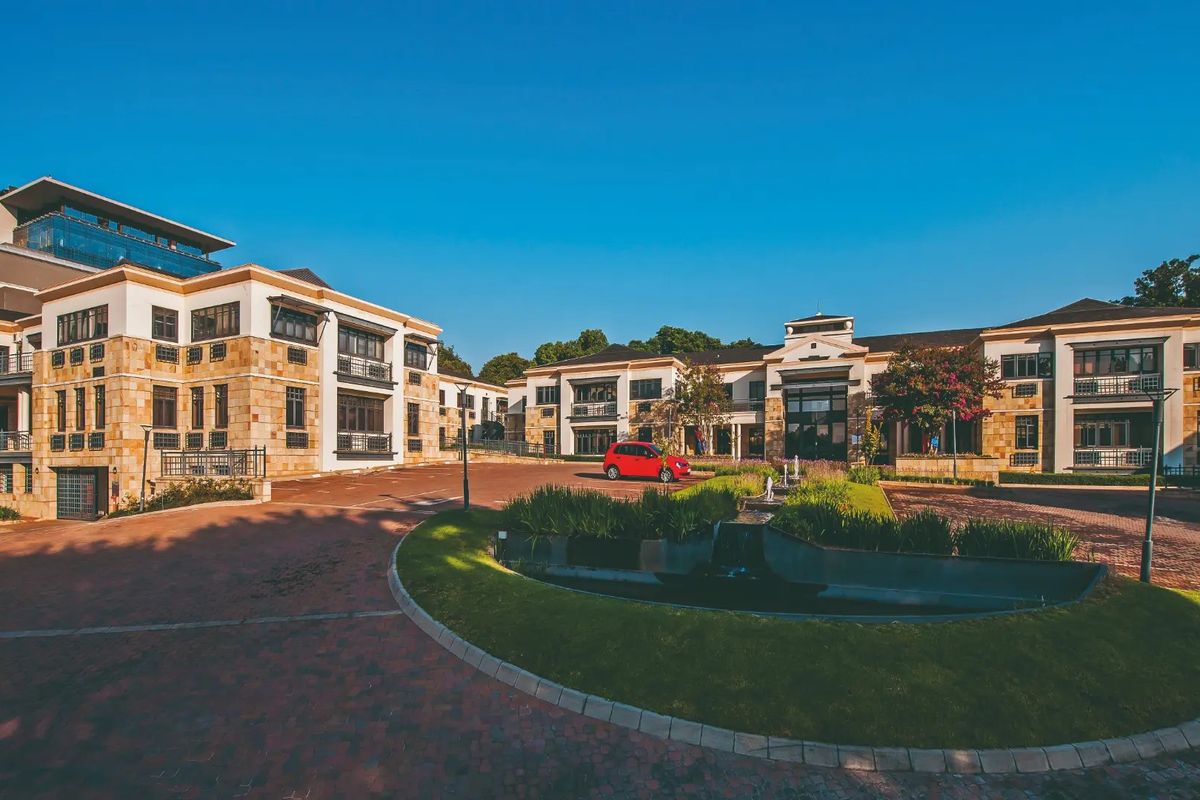 A professional exterior view of a multi-story commercial office complex featuring stone cladding, paved driveways, and a central landscaped water feature.