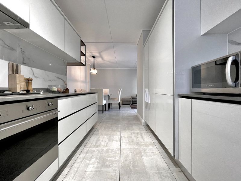 Modern galley-style kitchen featuring high-gloss white cabinetry, integrated appliances, marble-effect splashback, and sleek stone-look floor tiles leading into the dining area.