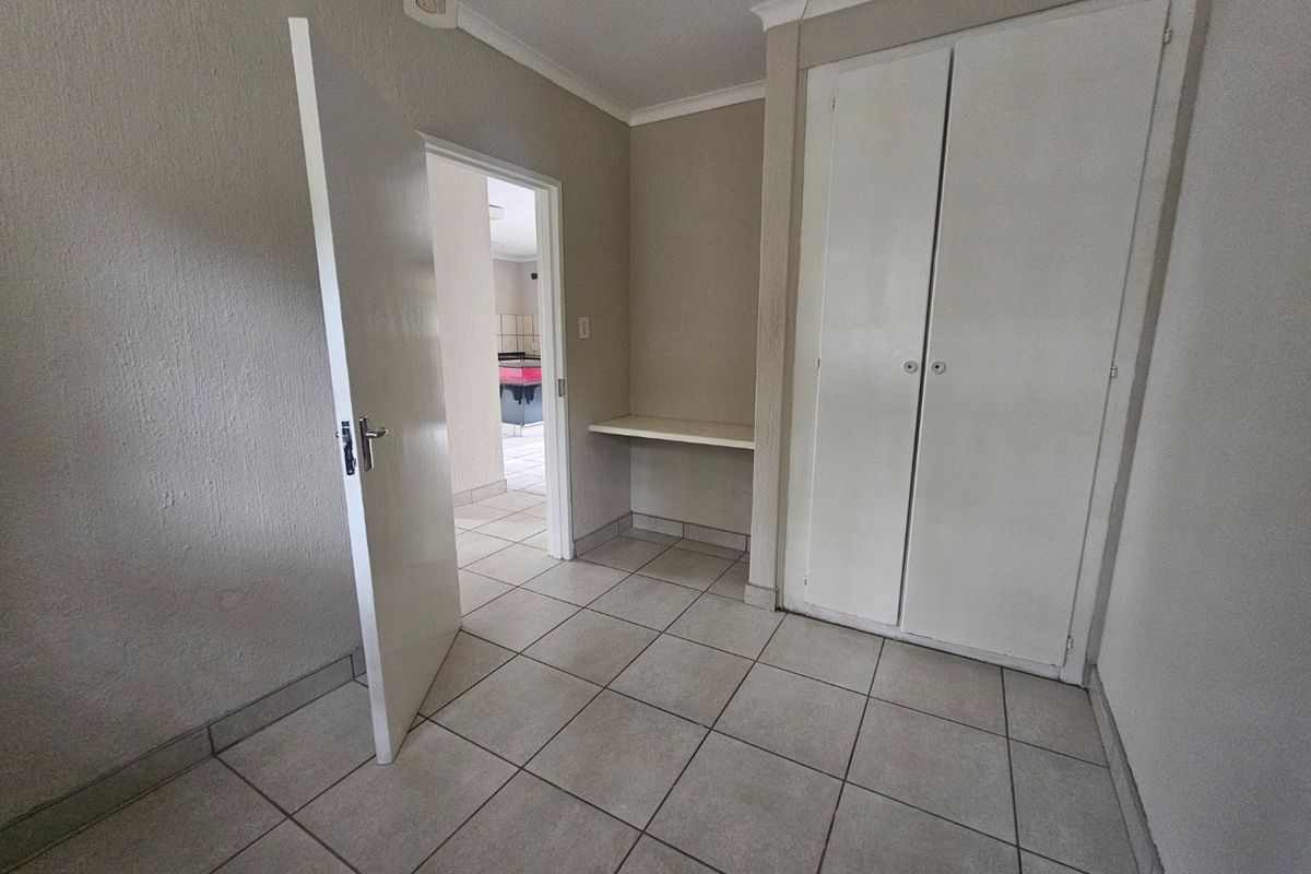 A clean, empty bedroom featuring neutral tiled flooring, a built-in wardrobe, and a convenient built-in desk area with access to the adjacent room.