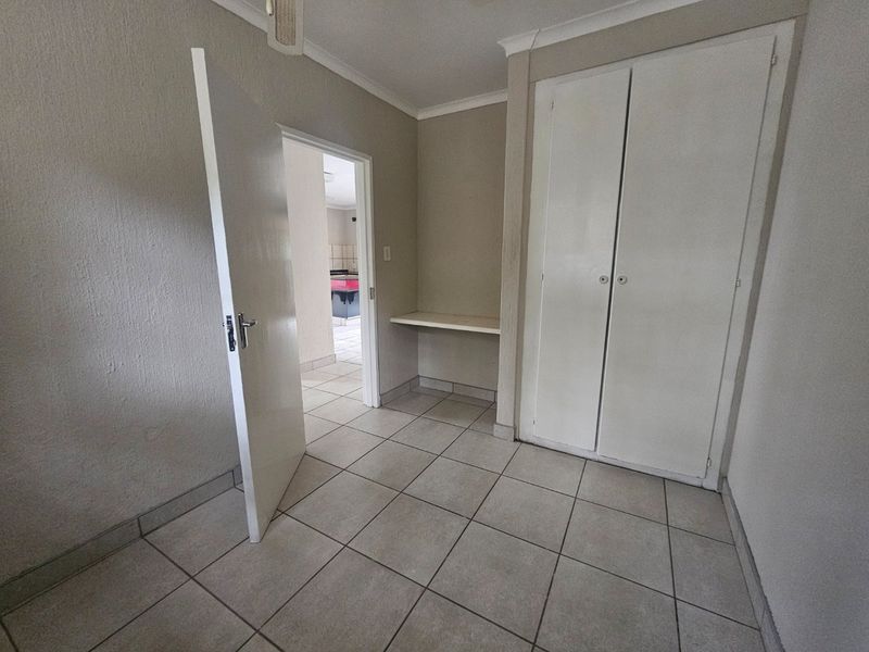 A clean, empty bedroom featuring neutral tiled flooring, a built-in wardrobe, and a convenient built-in desk area with access to the adjacent room.
