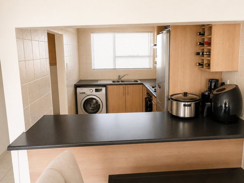 Functional kitchen featuring light wood cabinetry, dark countertops, integrated washing machine, and a convenient breakfast bar pass-through to the living area.