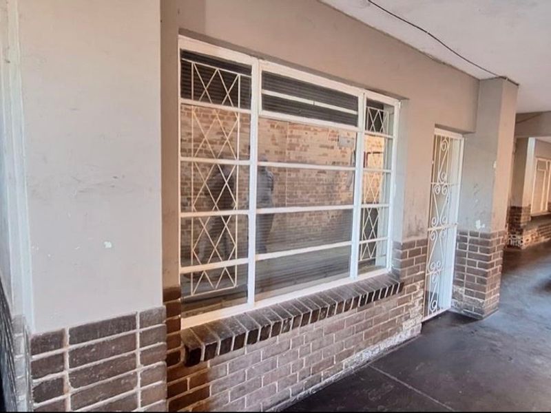 A covered exterior hallway or walkway featuring brick accents, security gates, and metal-framed windows, showing signs of wear and requiring maintenance.