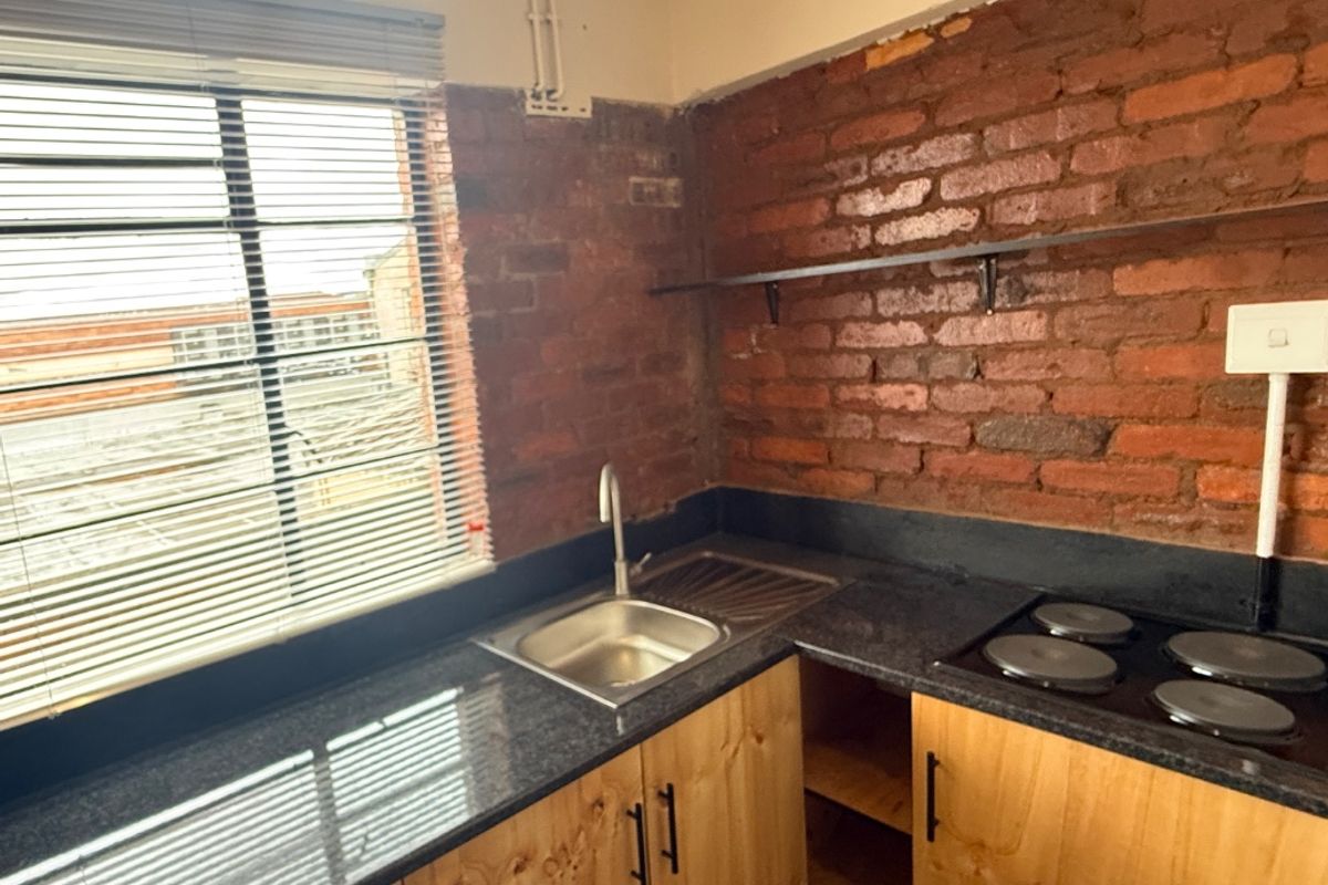 Compact kitchen featuring exposed brick walls, granite countertops, stainless steel sink, built-in electric hob, and custom plywood cabinetry.