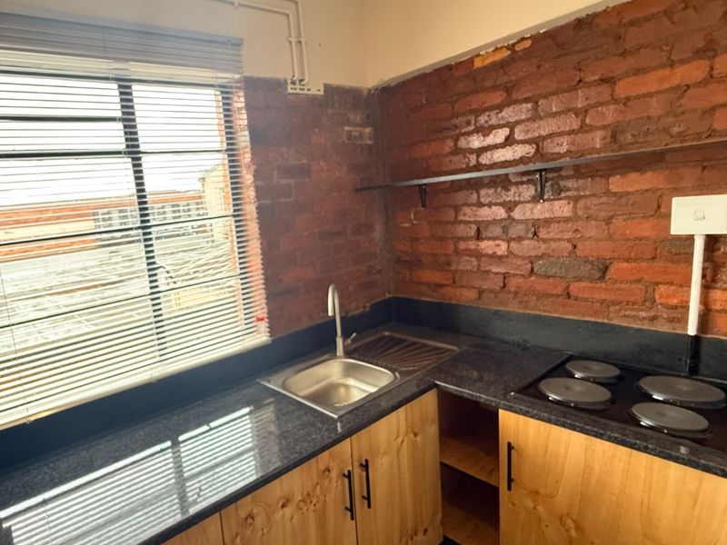 Compact kitchen featuring exposed brick walls, granite countertops, stainless steel sink, built-in electric hob, and custom plywood cabinetry.
