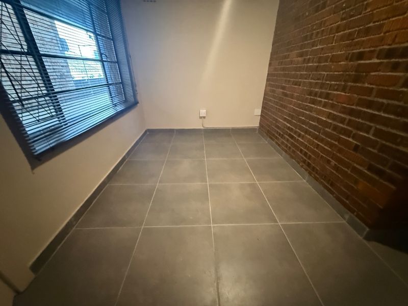 An empty living area featuring a rustic exposed brick wall, neutral tiled flooring, and venetian blinds on the window.