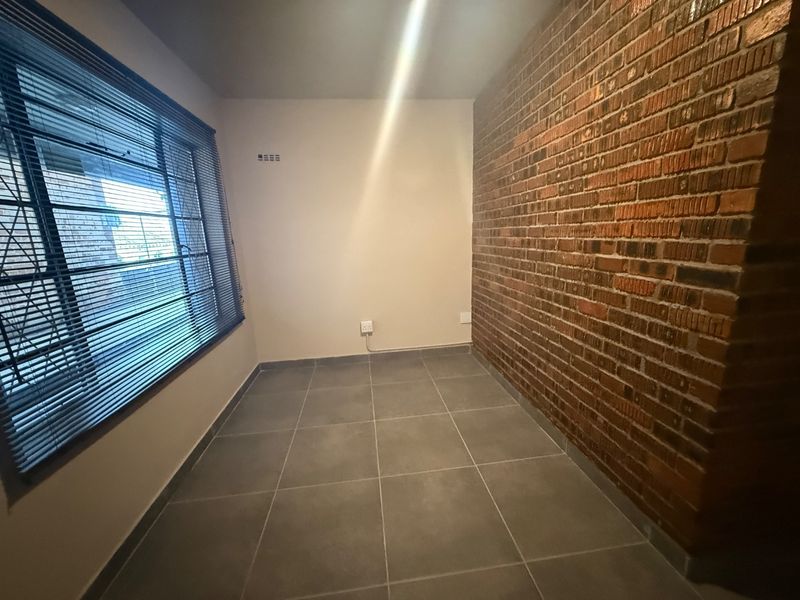 A clean, empty bedroom featuring a striking exposed brick feature wall, modern grey floor tiles, and large windows with fitted venetian blinds.