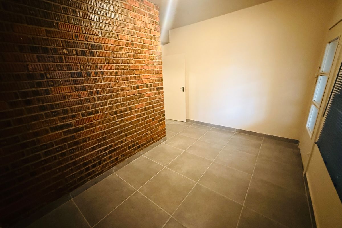 An empty living space featuring a prominent exposed brick wall, neutral tiled flooring, and natural light from a side window.