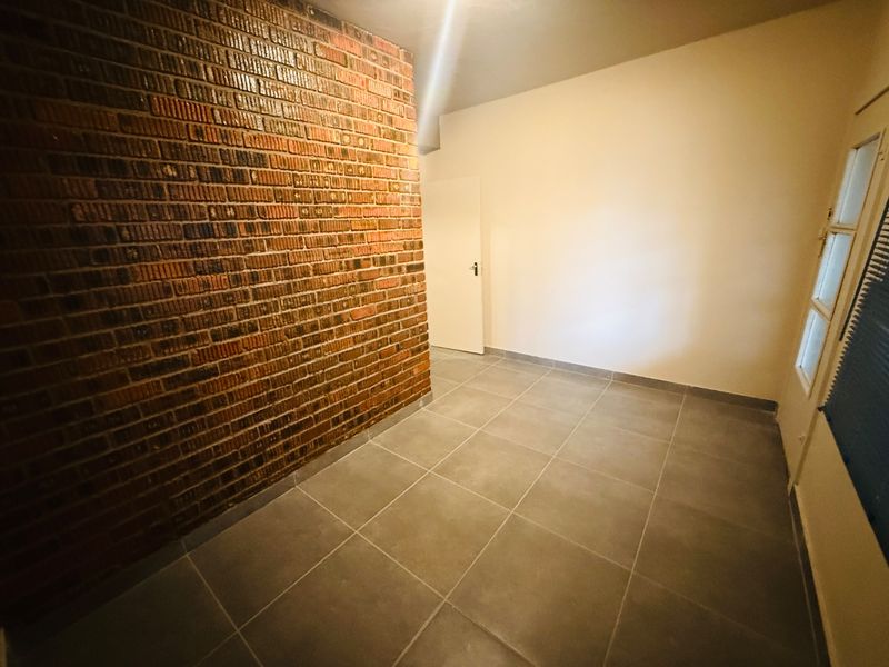 An empty living space featuring a prominent exposed brick wall, neutral tiled flooring, and natural light from a side window.