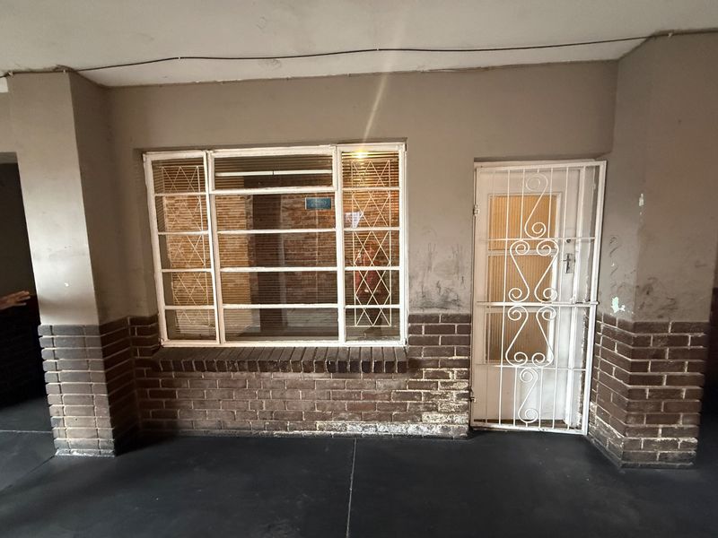 Entrance area featuring a security gate, brick facade detailing, and a steel-framed window, showing signs of wear and requiring cosmetic maintenance.