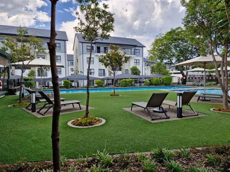 A well-maintained communal swimming pool area featuring landscaped gardens, paved lounging spaces, and modern apartment building architecture in the background.