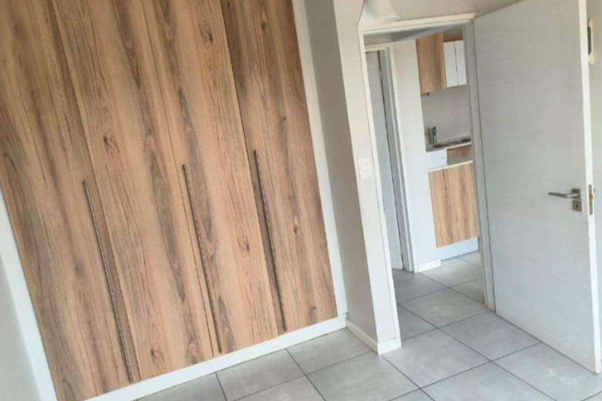 A clean, empty bedroom featuring modern wood-finish built-in cupboards and neutral tiled flooring, with a view into an adjacent kitchen area.