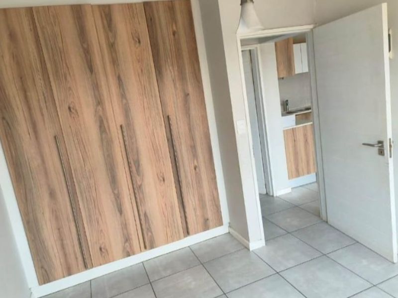 A clean, empty bedroom featuring modern wood-finish built-in cupboards and neutral tiled flooring, with a view into an adjacent kitchen area.