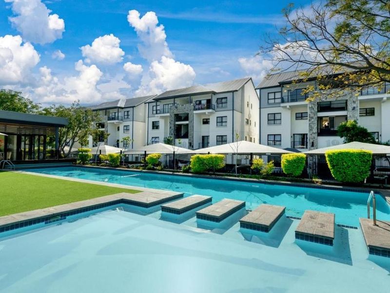 A well-maintained communal swimming pool area featuring a modern pool deck, landscaped garden, and views of the surrounding apartment complex buildings.