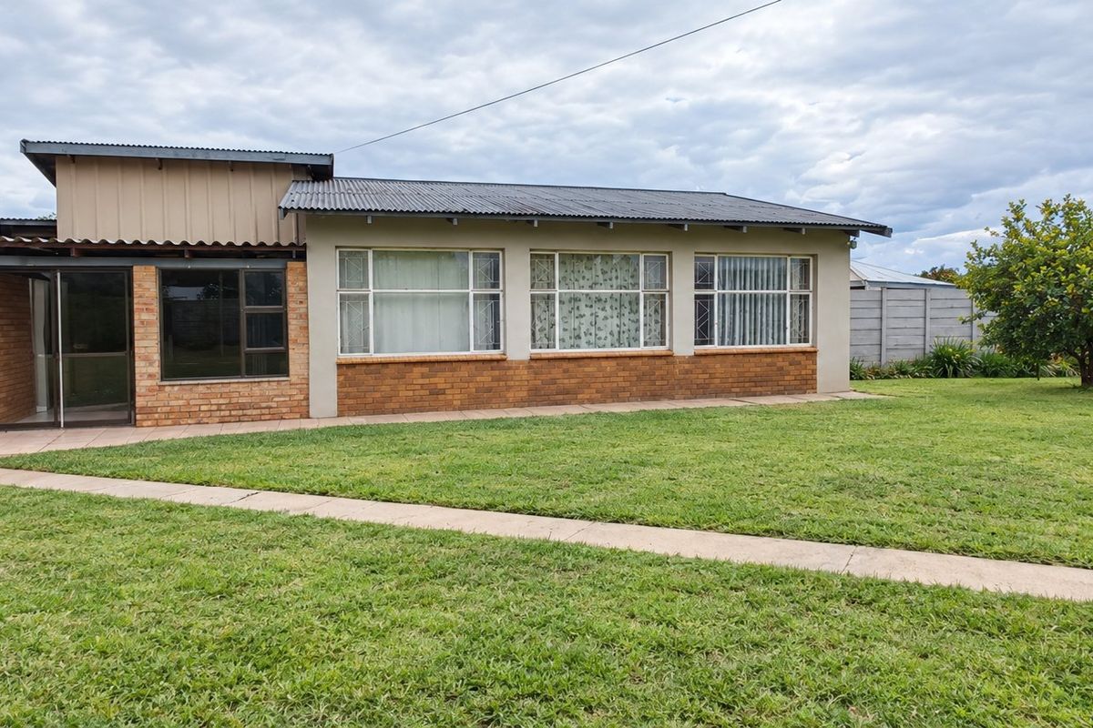Rear view of a single-story brick house featuring a corrugated metal roof, large windows, a paved walkway, and an attached carport set in a garden.