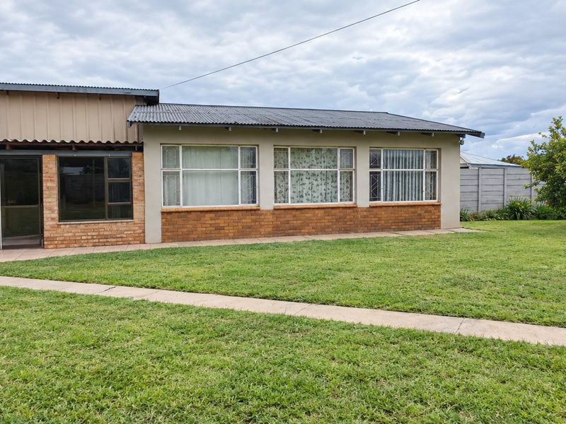 Rear view of a single-story brick house featuring a corrugated metal roof, large windows, a paved walkway, and an attached carport set in a garden.