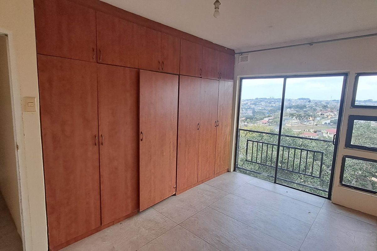 A spacious, empty bedroom featuring extensive built-in wooden cupboards, tiled flooring, and a sliding glass door leading to a balcony with scenic views.