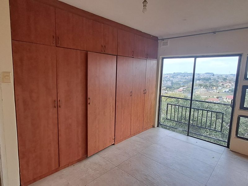 A spacious, empty bedroom featuring extensive built-in wooden cupboards, tiled flooring, and a sliding glass door leading to a balcony with scenic views.