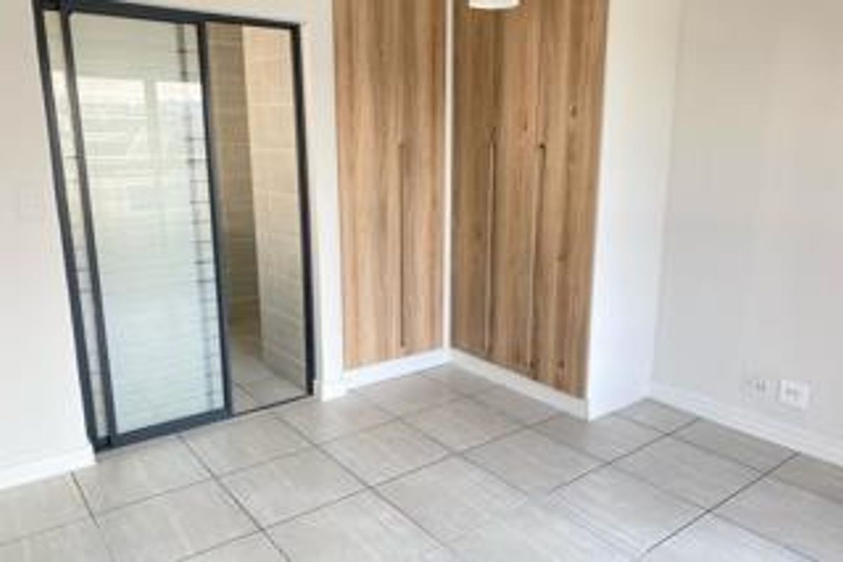 A bright, modern bedroom featuring neutral tiled flooring, built-in wooden cupboards, and a sliding glass door leading to an adjacent space.
