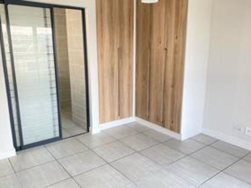 A bright, modern bedroom featuring neutral tiled flooring, built-in wooden cupboards, and a sliding glass door leading to an adjacent space.