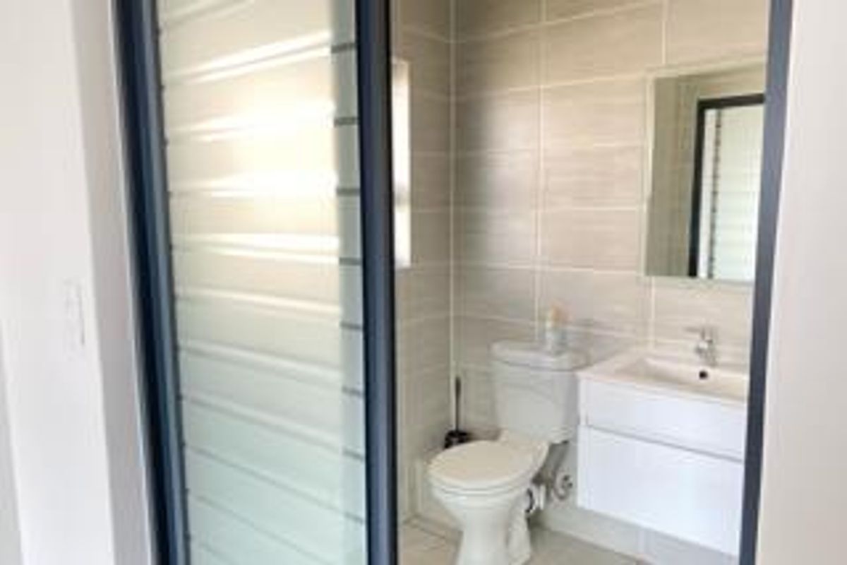 A modern, clean bathroom featuring a wall-mounted vanity, ceramic toilet, and neutral wall tiling, accessed via a sleek sliding glass door.