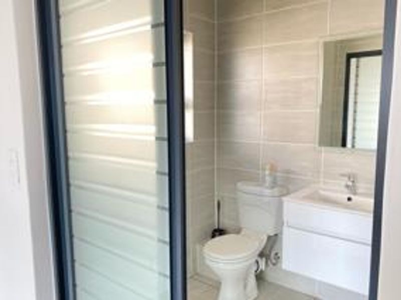 A modern, clean bathroom featuring a wall-mounted vanity, ceramic toilet, and neutral wall tiling, accessed via a sleek sliding glass door.