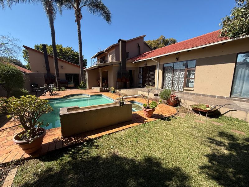 Rear exterior view featuring a swimming pool, paved patio area, built-in braai, and established garden with mature palm trees.