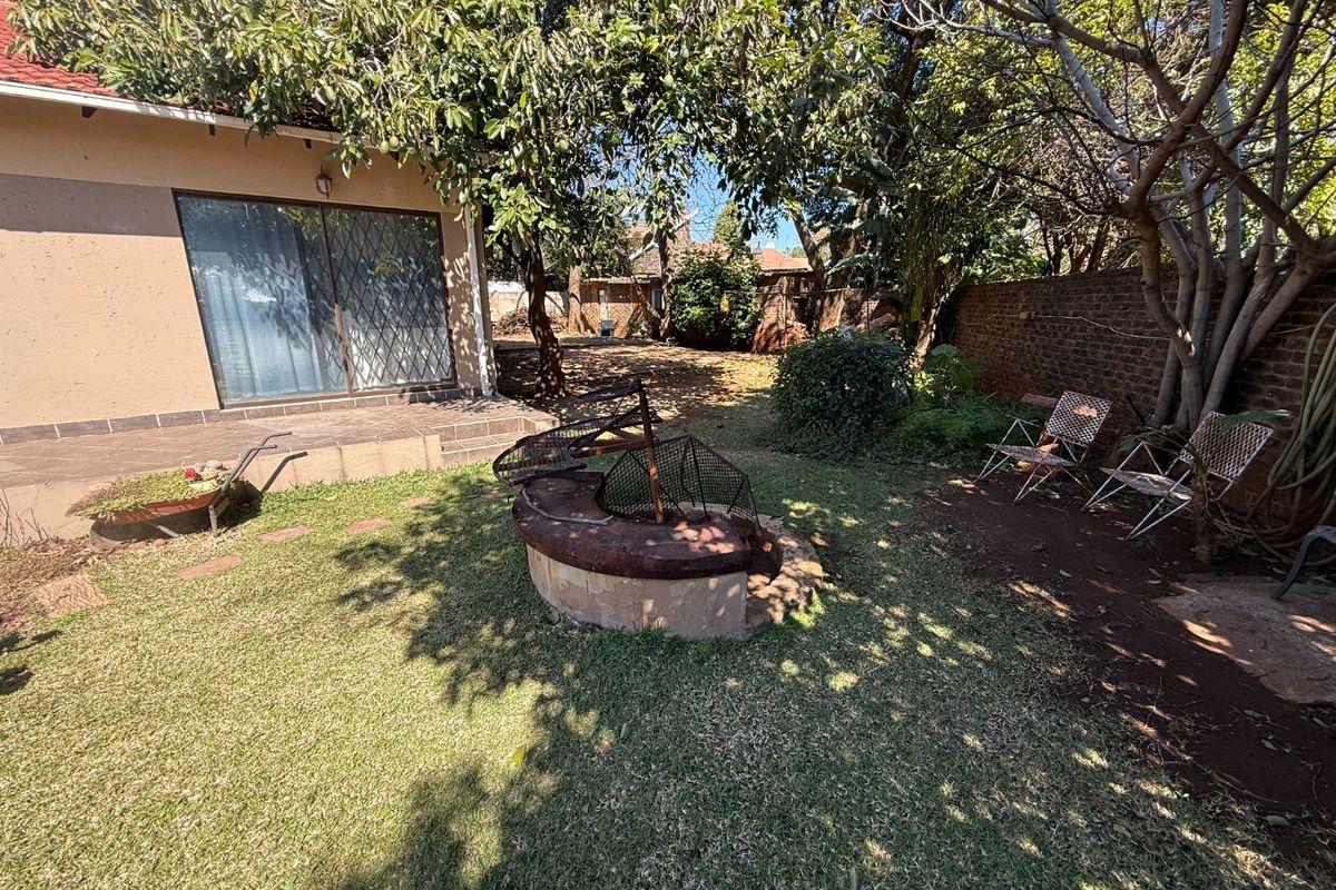 Rear garden area featuring a lawn, mature trees, a built-in braai, and a tiled patio area accessible via sliding glass doors.