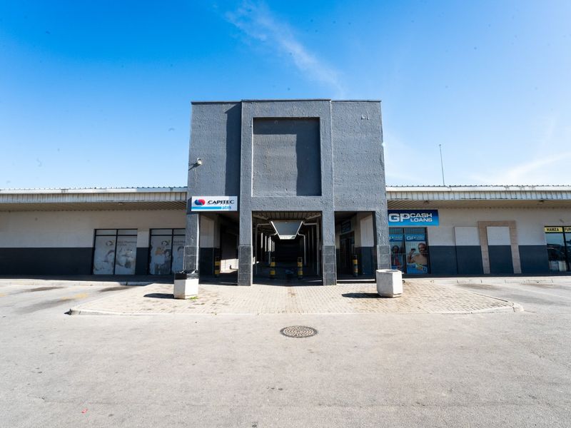 Front elevation of a commercial retail center featuring a central covered walkway, paved parking area, and multiple storefronts with signage.