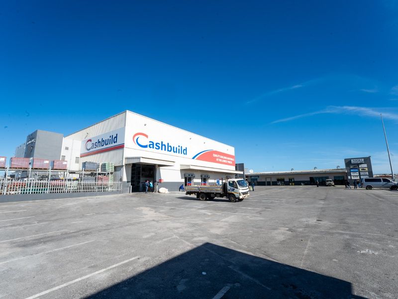 Wide-angle view of a commercial retail building featuring a large asphalt parking area, metal facade, and multiple roller shutter access points.