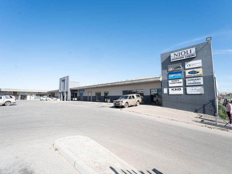 Exterior view of Njoli Shopping Centre featuring a large asphalt parking area and multiple commercial tenant signs on the building facade.