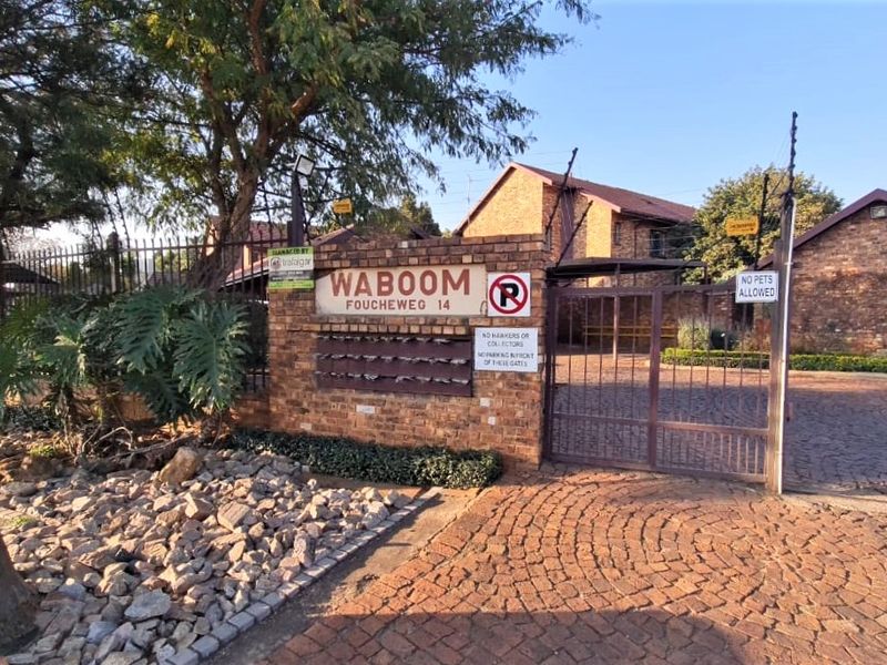 The entrance to the Waboom apartment complex featuring a brick facade, secure metal gate, electric fencing, and paved driveway access.