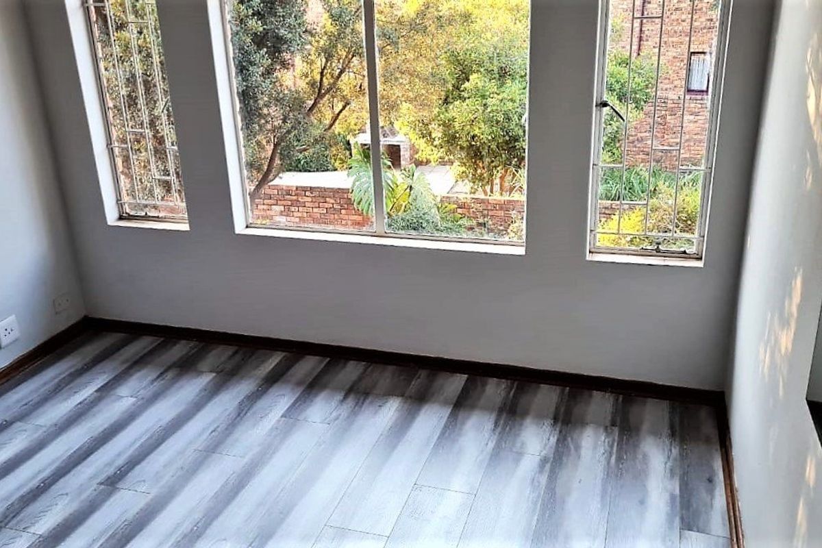 A bright, empty bedroom featuring modern grey-toned laminate flooring and large windows with security bars overlooking the garden.