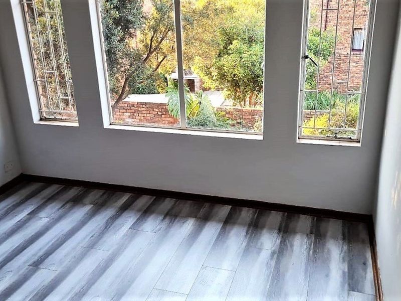 A bright, empty bedroom featuring modern grey-toned laminate flooring and large windows with security bars overlooking the garden.