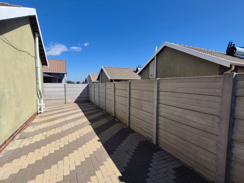A side exterior view of the property featuring paved ground surfaces and a precast concrete boundary wall under a clear sky.