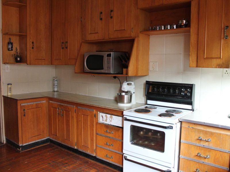 A dated kitchen featuring original wooden cabinetry, laminate countertops, tiled backsplash, and a freestanding electric stove with hardwood flooring.