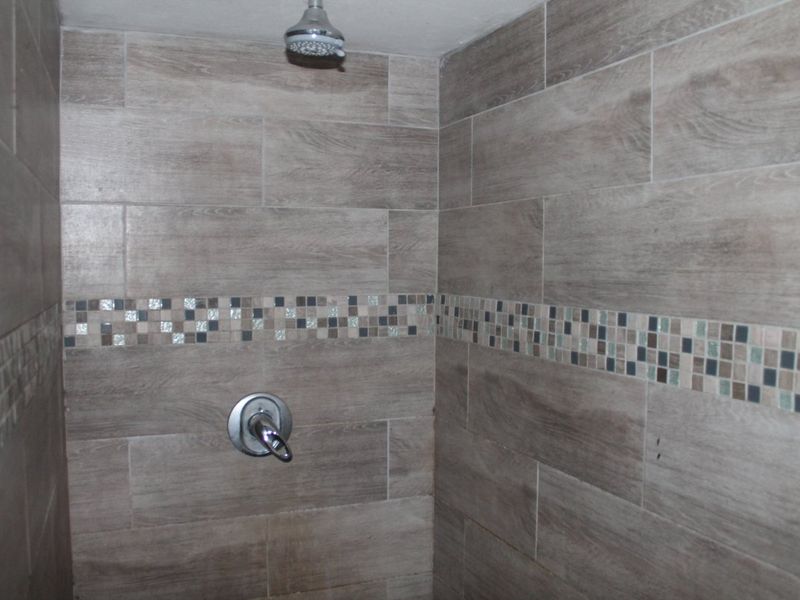 A shower enclosure featuring wood-look ceramic wall tiles and a decorative mosaic accent border with a standard shower mixer and head.