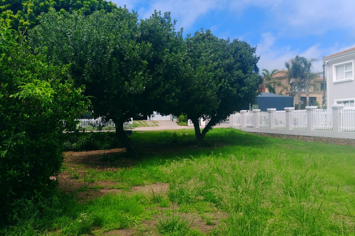 A view of a residential garden plot featuring mature trees, established lawn, and a white picket fence boundary.