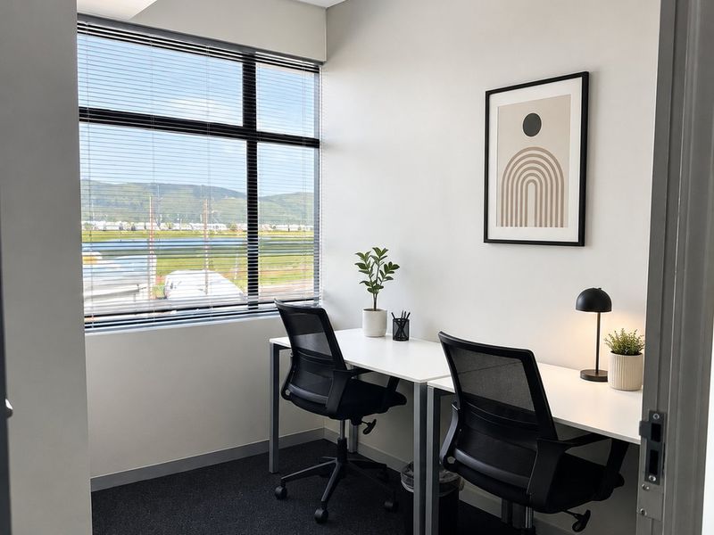 A modern, well-lit office space featuring large windows with venetian blinds, neutral walls, and dark carpeting, suitable for a professional work environment.