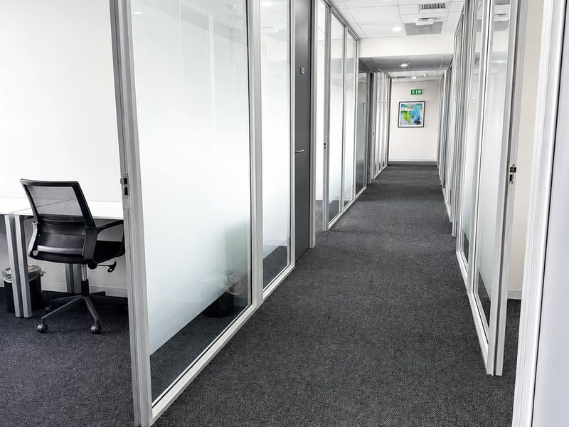 A modern commercial office hallway featuring glass partition walls, carpeted flooring, and a suspended ceiling with integrated lighting and air conditioning.