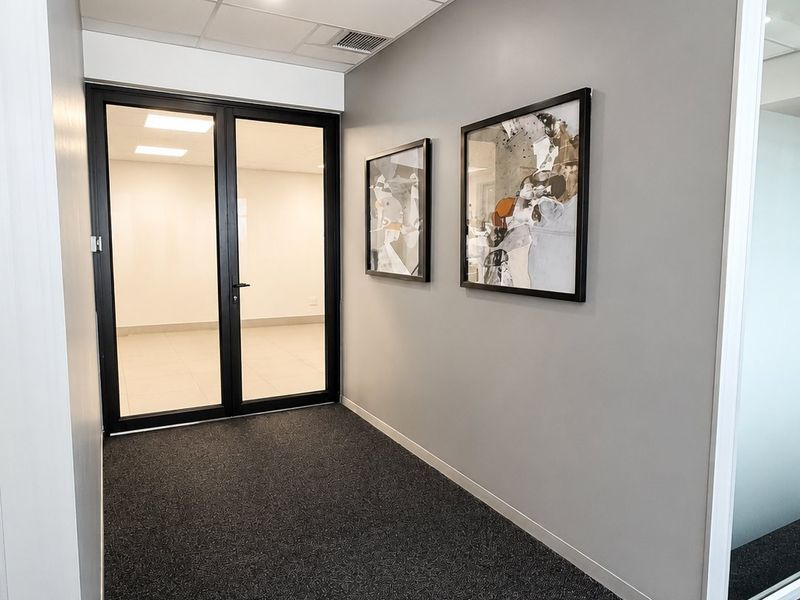 A clean, modern commercial hallway featuring dark carpet flooring, grey walls, and black-framed glass double doors leading into an adjacent office space.