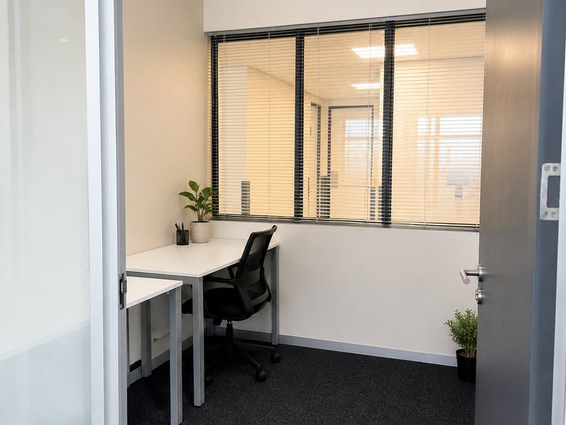 A compact, modern office space featuring carpeted flooring, recessed ceiling lighting, and an internal window with fitted blinds for privacy.
