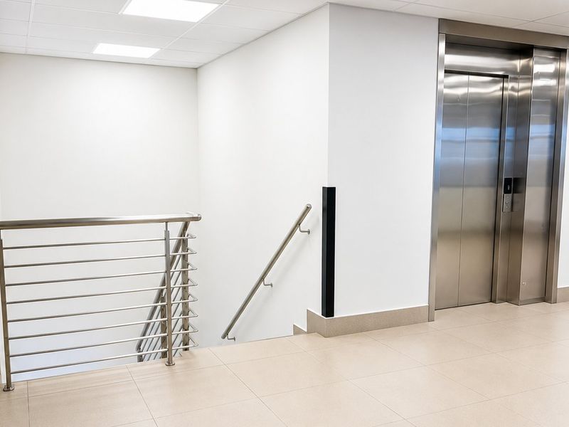Modern commercial hallway featuring a stainless steel elevator, matching balustrade, clean tiled flooring, and recessed ceiling lighting.