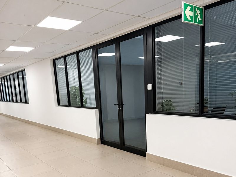 A modern commercial hallway featuring ceramic tiled flooring, suspended ceiling with recessed lighting, and black aluminium framed glass partitions leading to office spaces.