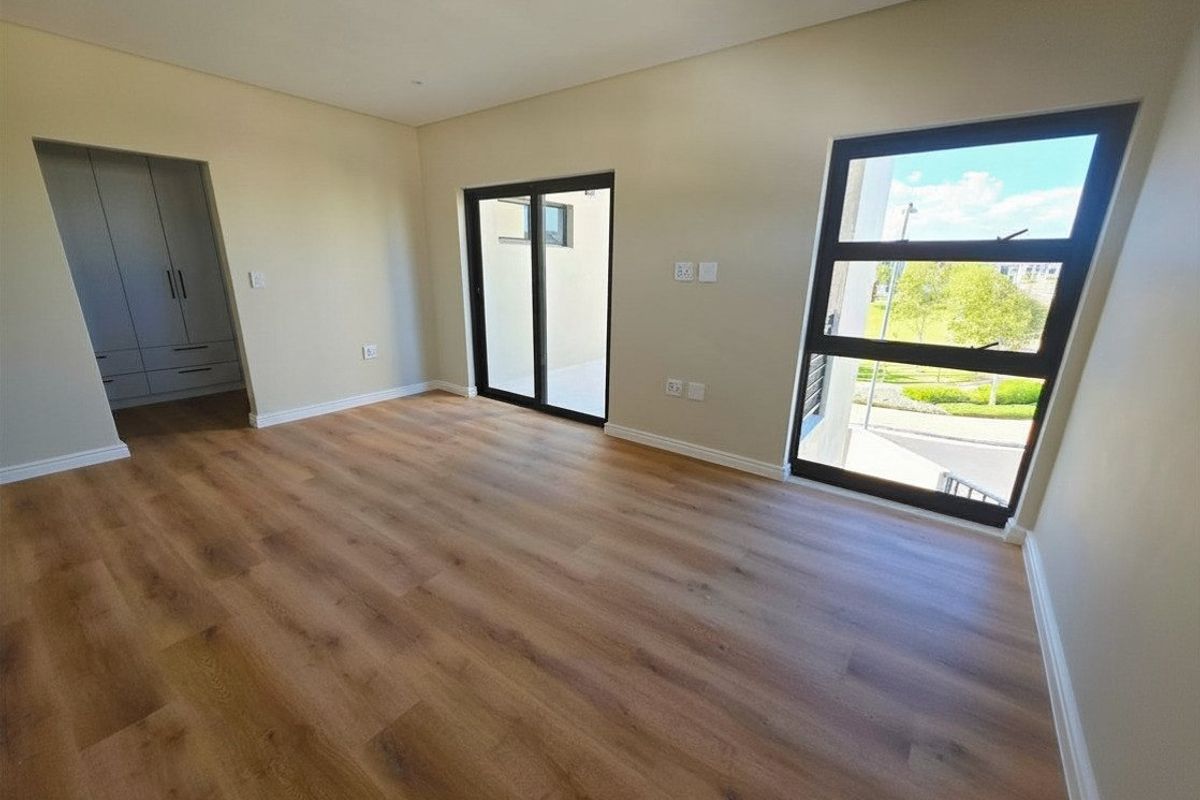 A bright, modern bedroom featuring laminate flooring, built-in cupboards, and large windows providing natural light and outdoor views.